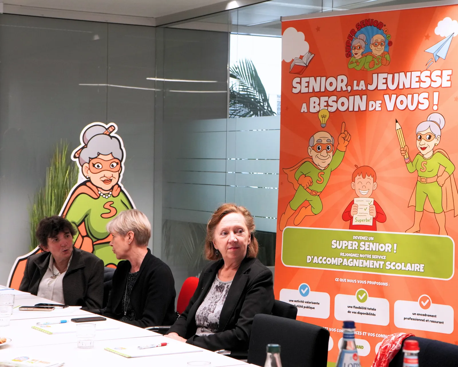 Participants autour de la table devant l’affiche « Senior, la jeunesse a besoin de vous ! » lors de la visite du Ministre Max Hahn au service Super Senior du MEC asbl