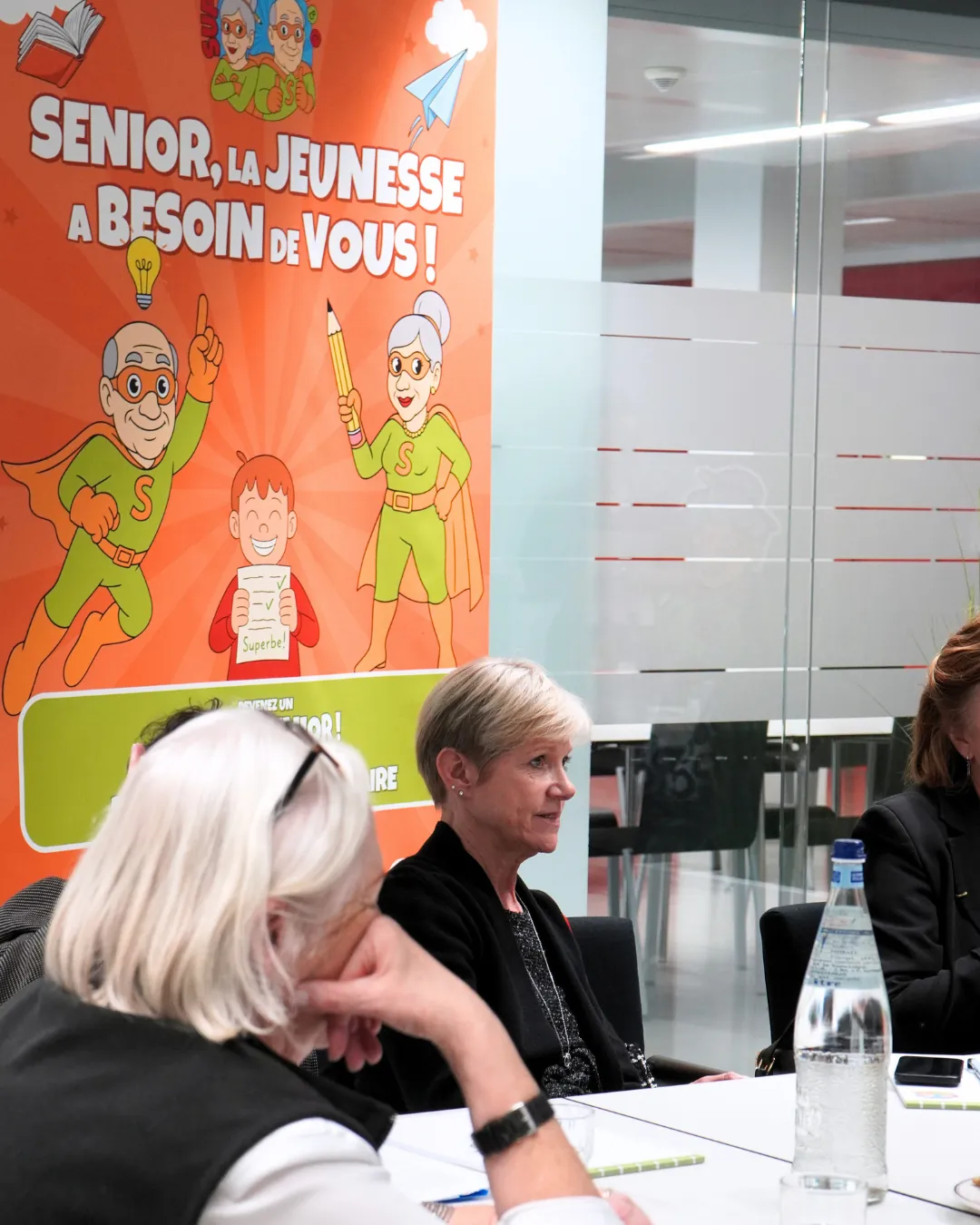 Échange entre participantes devant l’affiche de recrutement Super Senior lors de la visite du Ministre Max Hahn au service Super Senior du MEC asbl