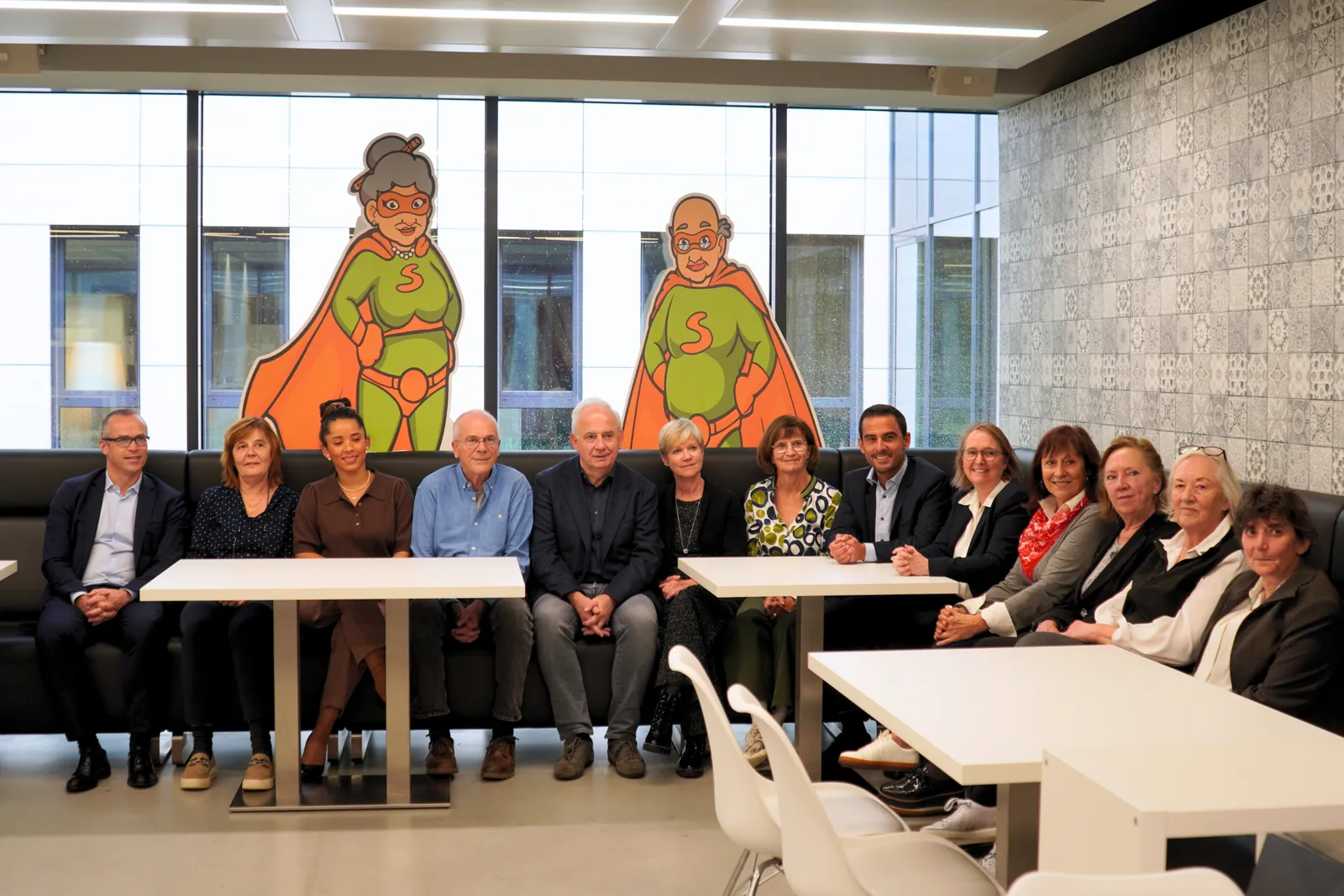 Photo de groupe des bénévoles et invités avec les mascottes Super Senior lors de la visite du Ministre Max Hahn au service Super Senior du MEC asbl