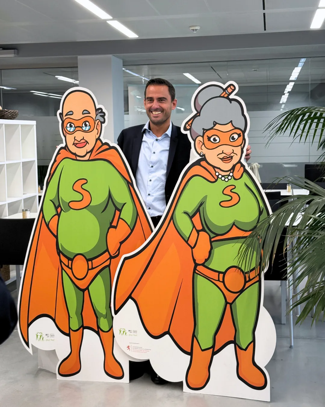 Invité posant entre les deux mascottes Super Senior lors de la visite du Ministre Max Hahn au service Super Senior du MEC asbl